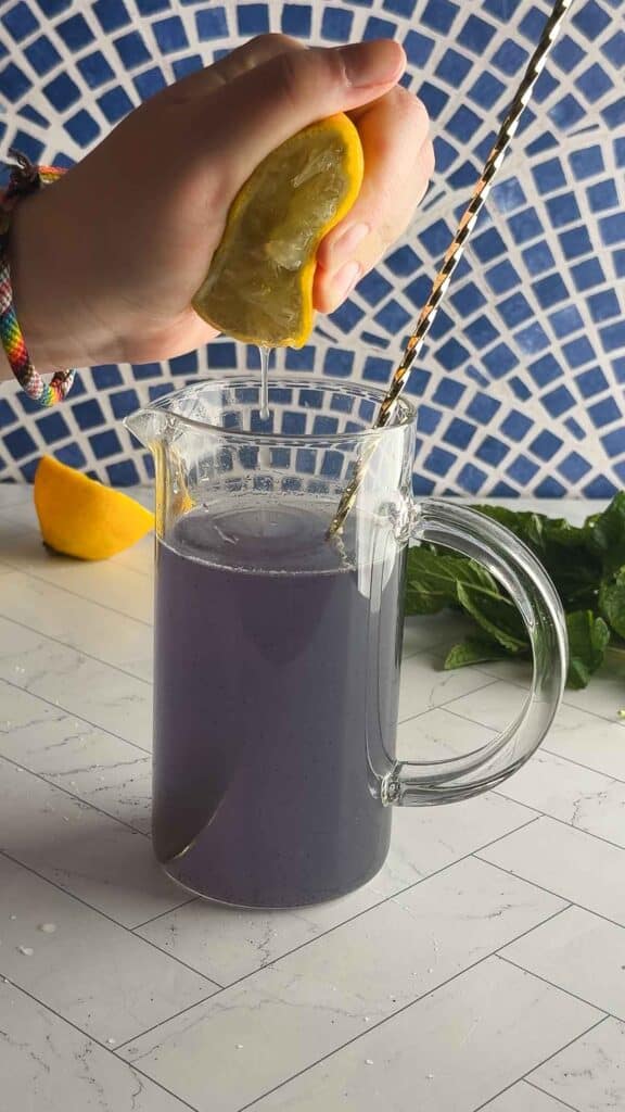 A hand squeezes a lemon over a pitcher of vibrant purple liquid with a straw on a white tiled surface, creating a refreshing Purple Electrolyte Mocktail.