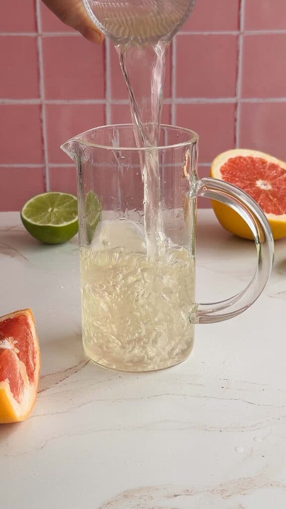 A clear liquid is being poured into a glass pitcher beside cut lime and grapefruit on a light countertop, perfect for a refreshing electrolyte mocktail recipe.