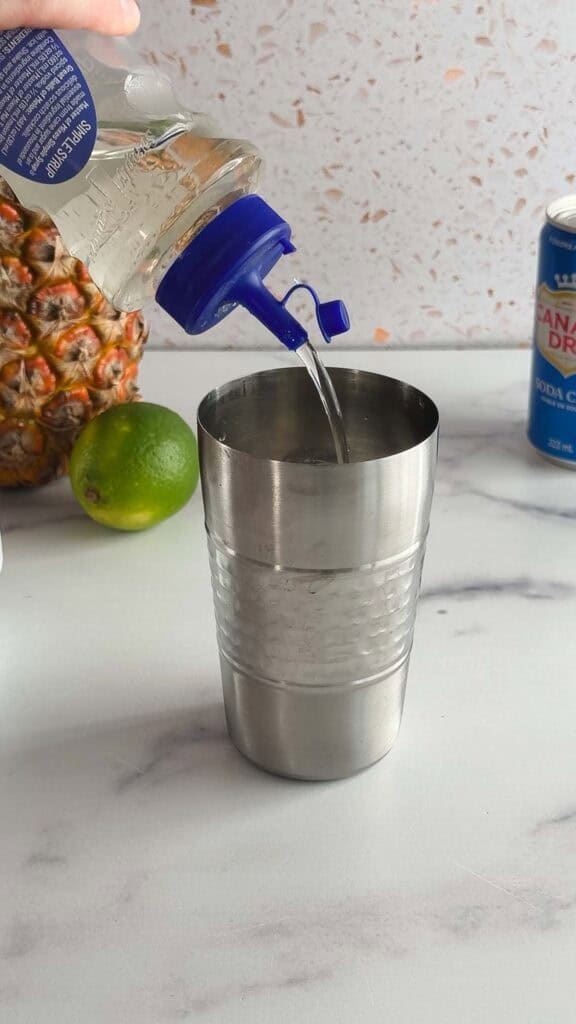 A hand pours clear liquid from a bottle into a metal shaker on a counter with a lime, pineapple, and soda can&mdash;perfect for crafting a refreshing tropical mocktail.