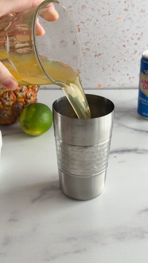 A hand pours juice from a glass into a metal cocktail shaker on a kitchen counter, crafting a refreshing tropical mocktail.