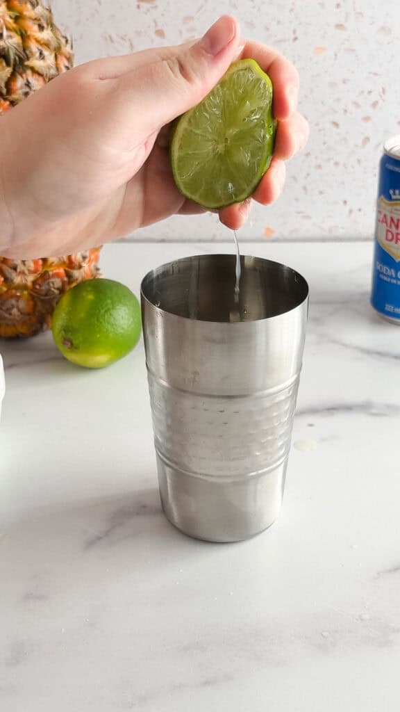 A hand squeezes a lime half over a metal cocktail shaker on a kitchen counter, preparing a refreshing tropical mocktail.