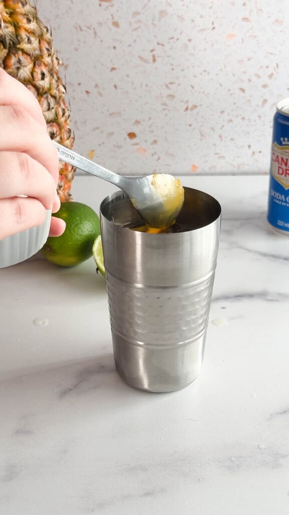 A hand holds a spoon, adding pineapple to a metal cup with limes and soda can in the background on a counter&mdash;the perfect setup for a refreshing Tropical Mocktail.