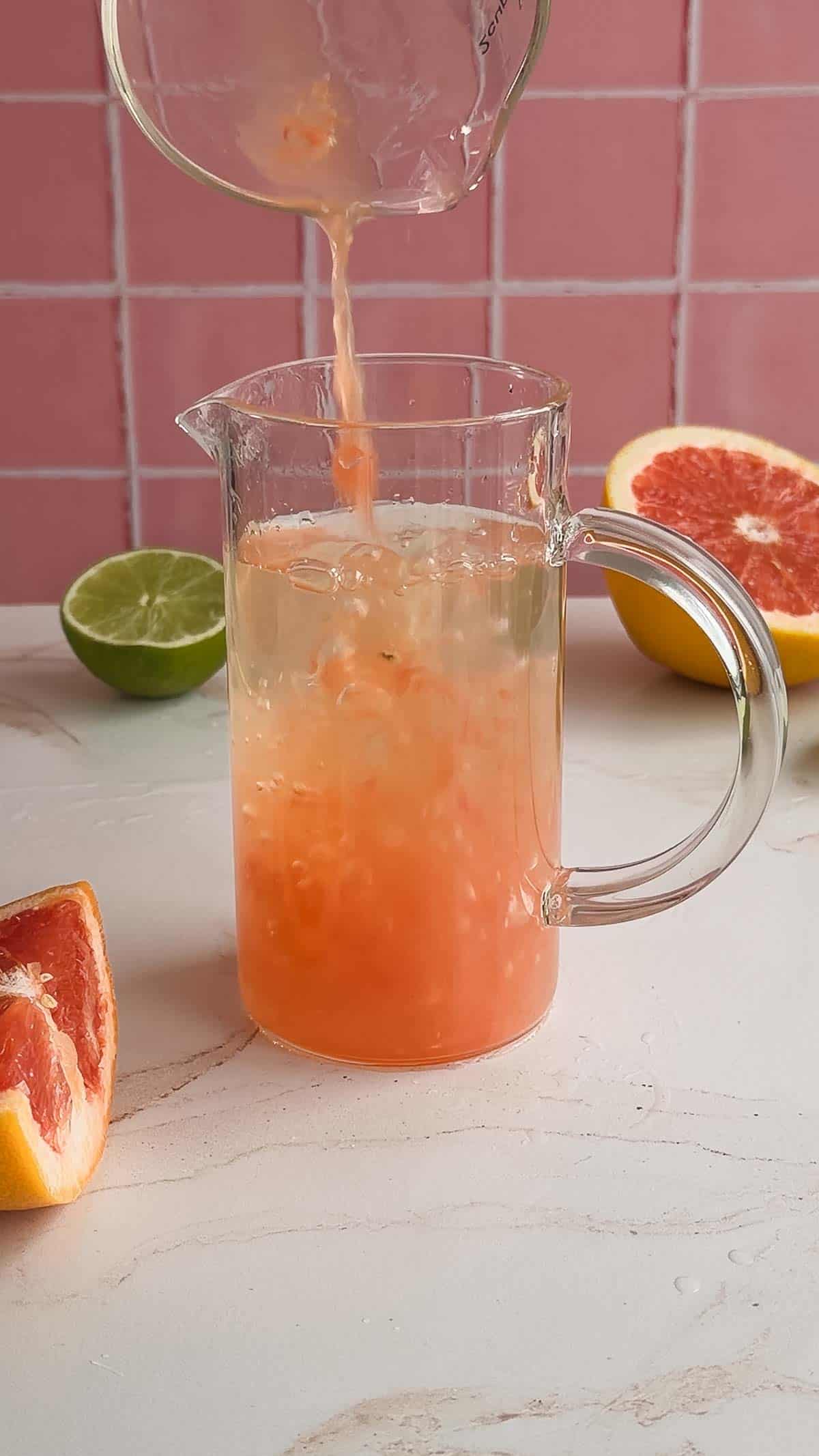 A glass pitcher with refreshing grapefruit juice being poured in, surrounded by cut grapefruit and lime on a white surface&mdash;perfect for crafting an energizing electrolyte mocktail recipe.
