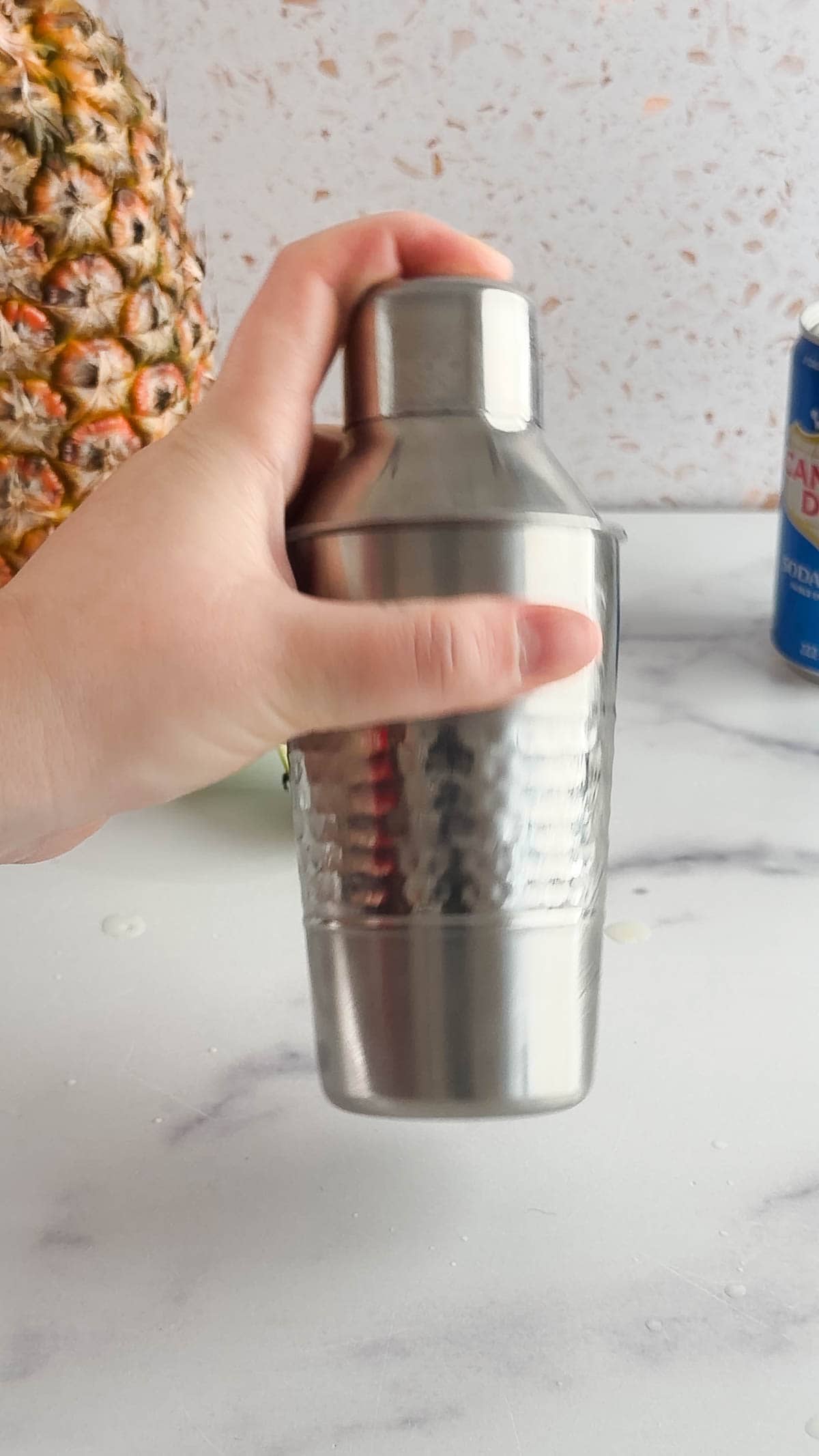 A hand holding a stainless steel cocktail shaker on a marble countertop with a pineapple and can nearby, ready to mix a refreshing tropical mocktail.