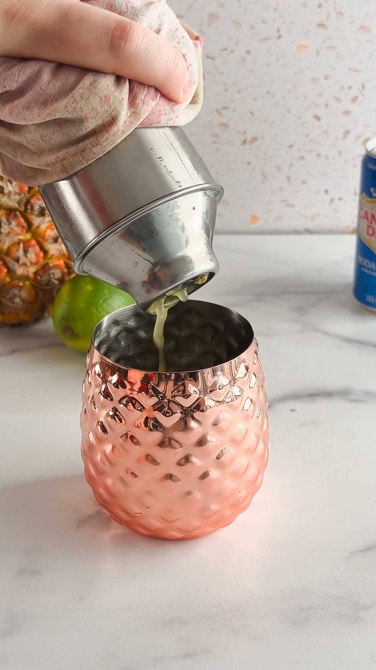 A tropical mocktail is being poured from a shaker into a textured copper cup on a marble counter.