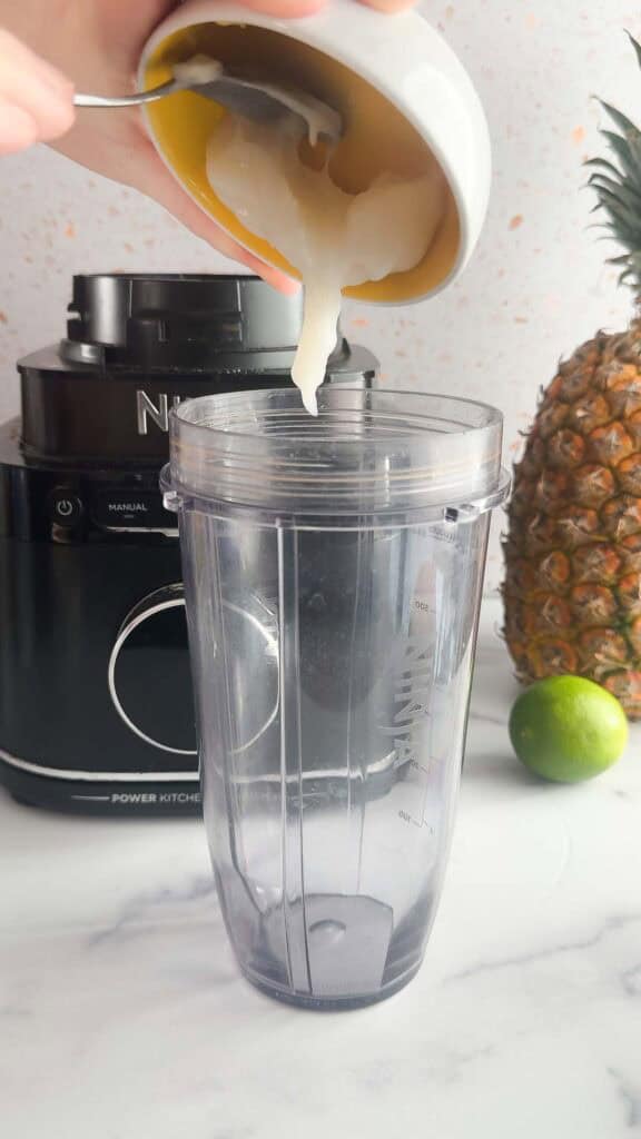 Yogurt being spooned into a blender cup, with a pineapple, lime, and blender in the background&mdash;perfect for creating a refreshing Mango Colada mocktail recipe.