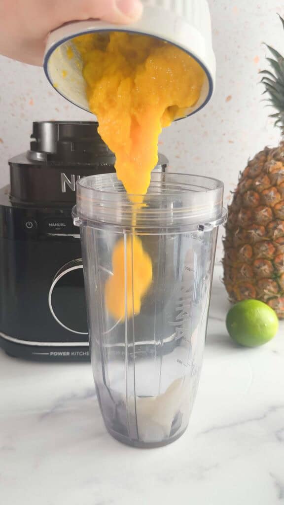Mango puree is poured into a blender cup, ready to create a refreshing Mango Colada. A pineapple, lime, and blender sit in the background, perfect for this vibrant mocktail recipe.