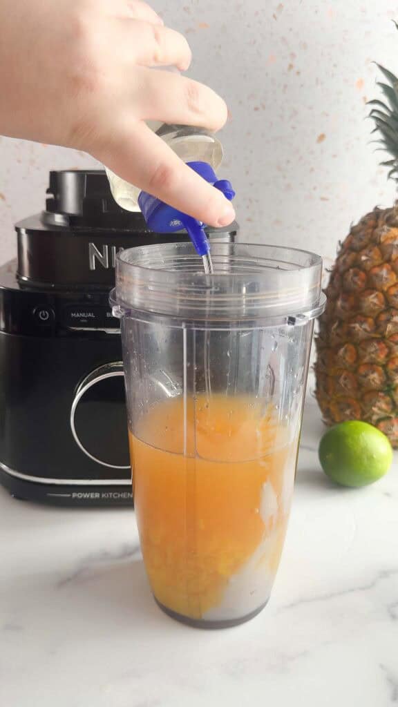 A hand pours liquid from a bottle into a blender cup with orange juice for a refreshing Mango Colada mocktail recipe, surrounded by lime, pineapple, and a blender.