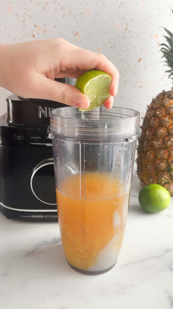 A hand squeezes a lime into a blender cup filled with orange liquid, preparing a refreshing Mango Colada mocktail recipe, with a blender and pineapple nearby.