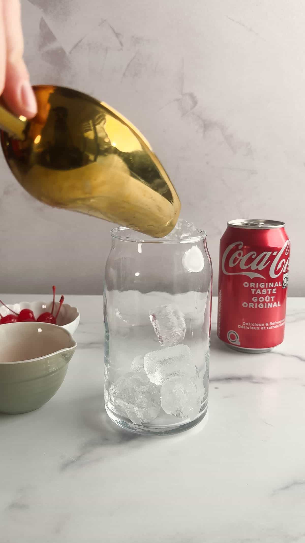 A gold scoop adds ice to a glass for a vintage Roy Rogers drink recipe, with a can of Coca-Cola and bowls of cherries on a white surface.