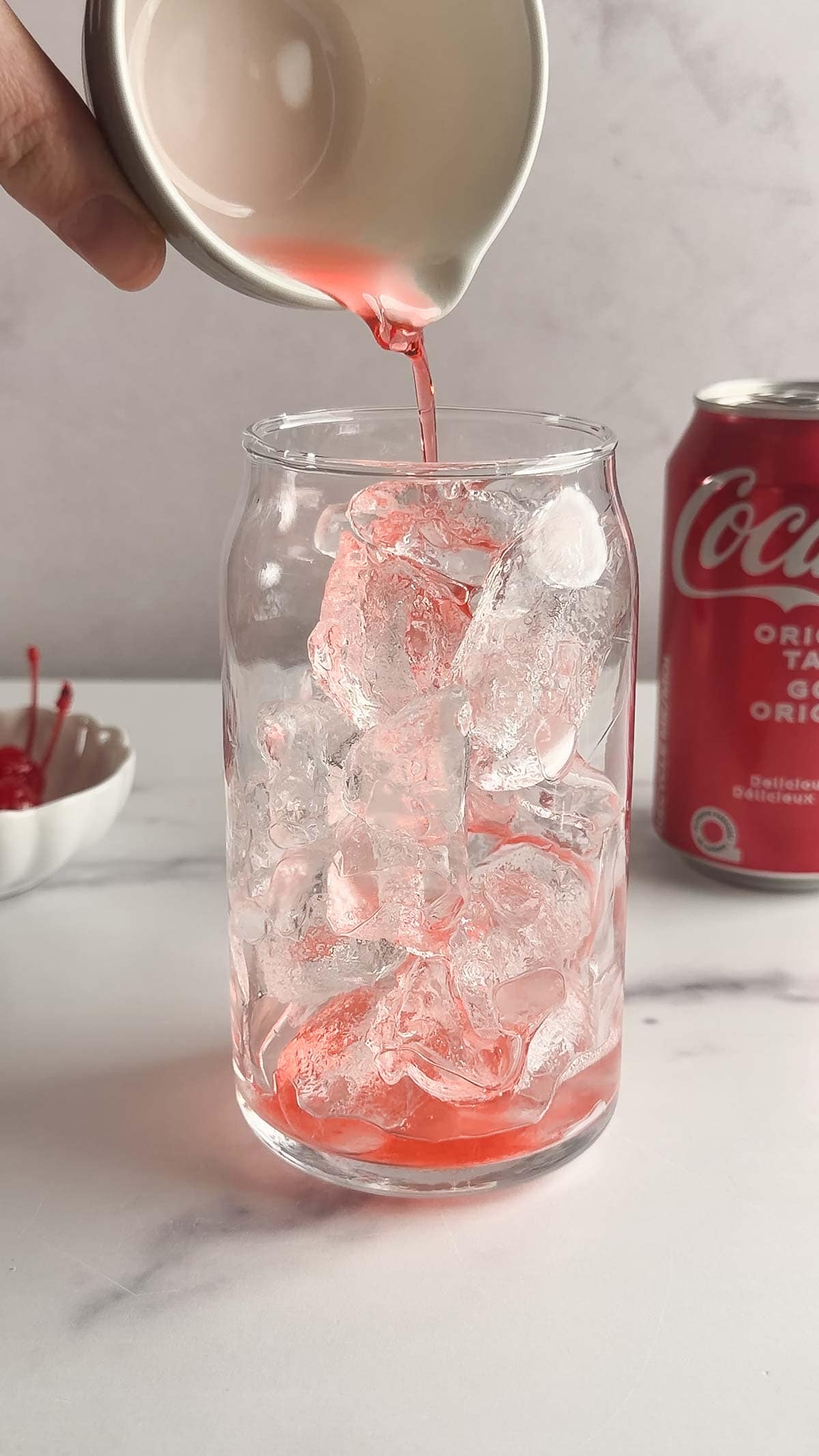 Pink syrup being poured over ice in a glass, with a can of Coca-Cola and cherries in the background&mdash;a nostalgic nod to the classic Roy Rogers recipe.