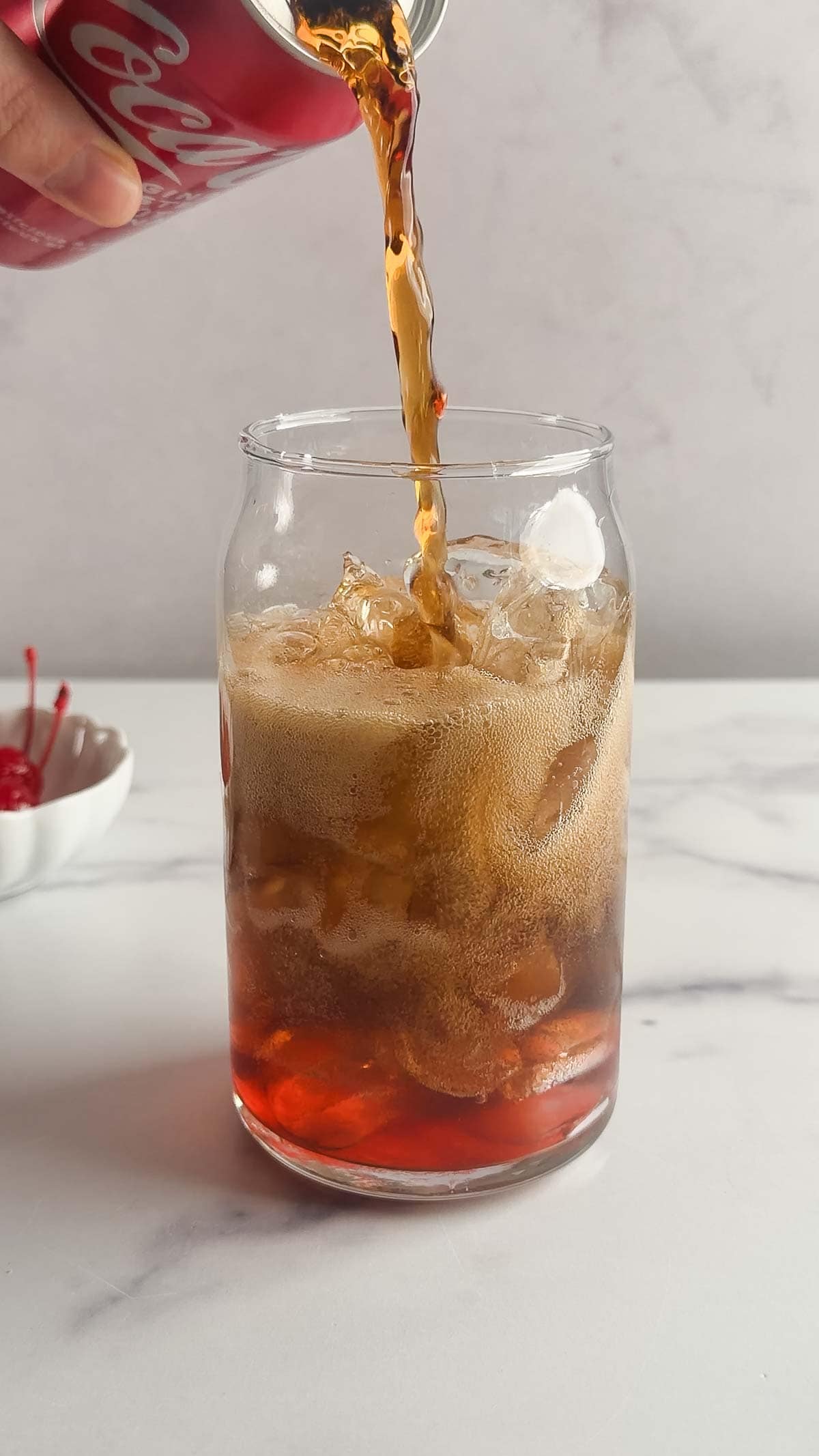 A hand pours cola from a can into a glass of ice on a white surface, with cherries in a dish nearby&mdash;classic ingredients for the vintage drink Roy Rogers.