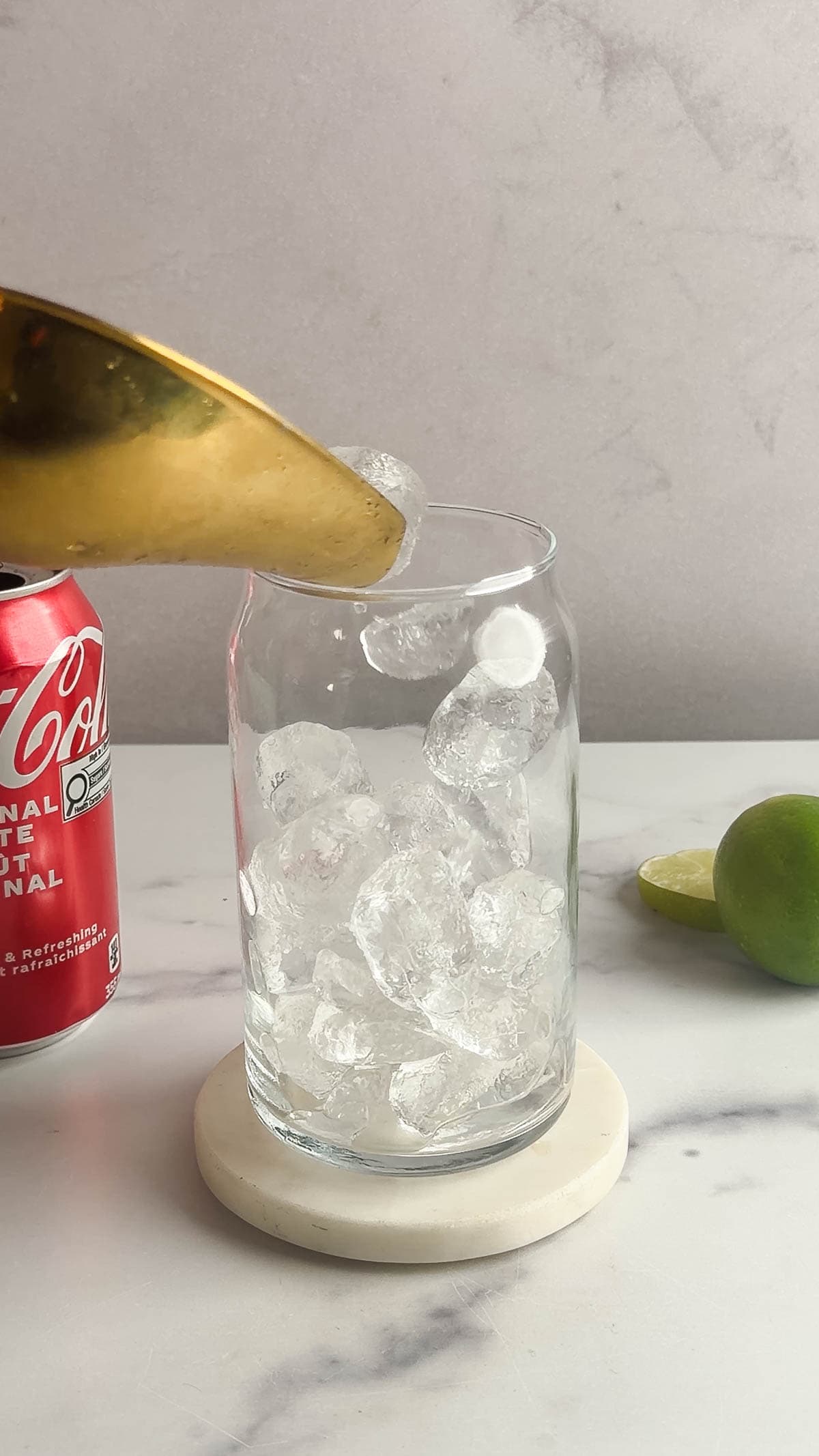 A glass is being filled with ice cubes from a scoop, with a soda can and limes nearby on the counter&mdash;perfect prep for a Dirty Soda mocktail recipe.