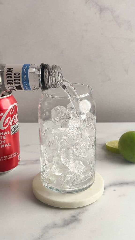 A clear coconut soda is being poured over ice in a glass, with a red cola can and lime halves nearby&mdash;perfect inspiration for your next mocktail recipe.