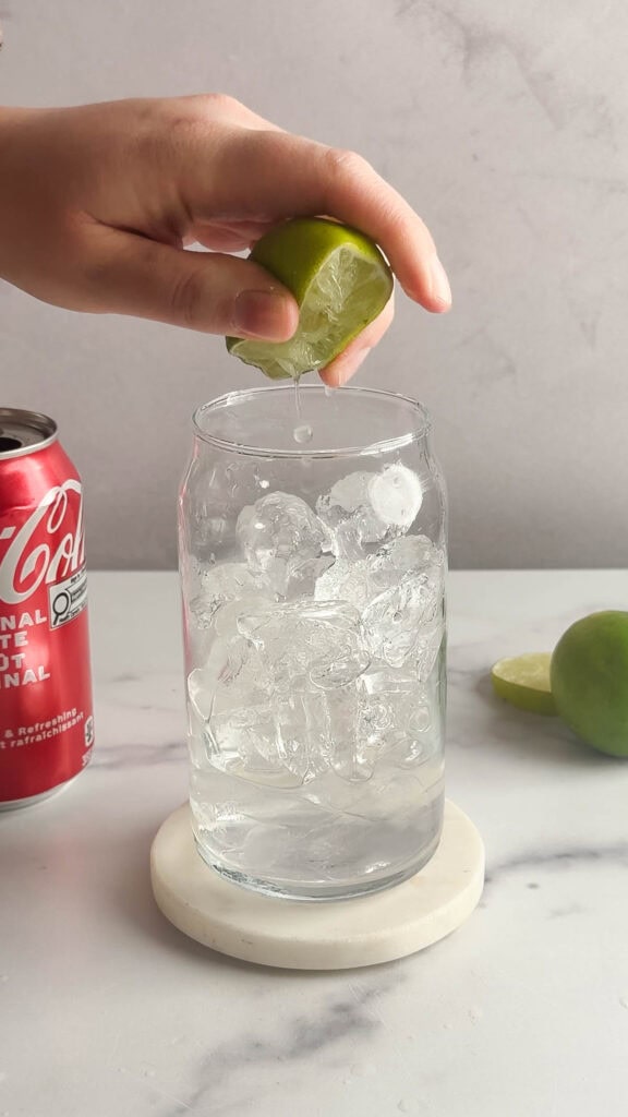 A hand squeezes a lime over a glass filled with ice, creating the perfect Dirty Soda mocktail recipe, with a can of Coke and lime halves nearby.