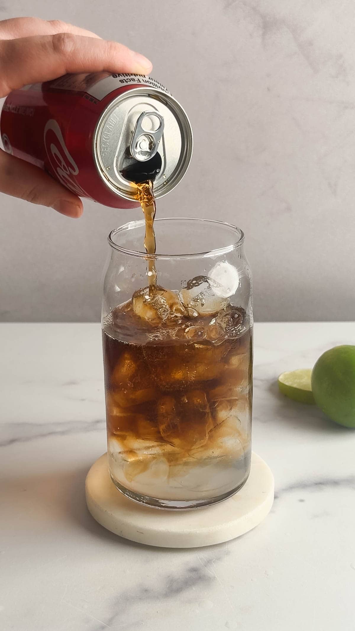 A hand pours cola from a can into a glass filled with ice on a marble surface, creating a refreshing Dirty Soda mocktail recipe, with lime nearby.