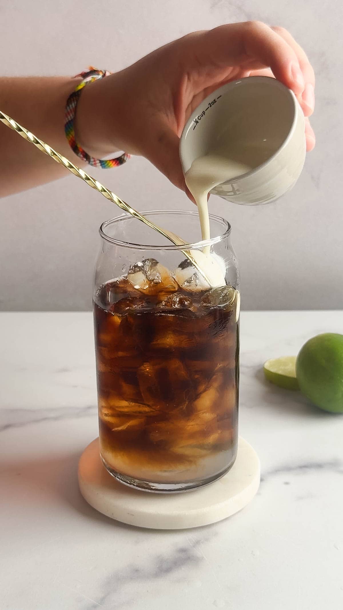 A hand pours cream into a glass of iced coffee; a lime and gold spoon are nearby on the marble surface, suggesting a refreshing coconut mocktail recipe twist.