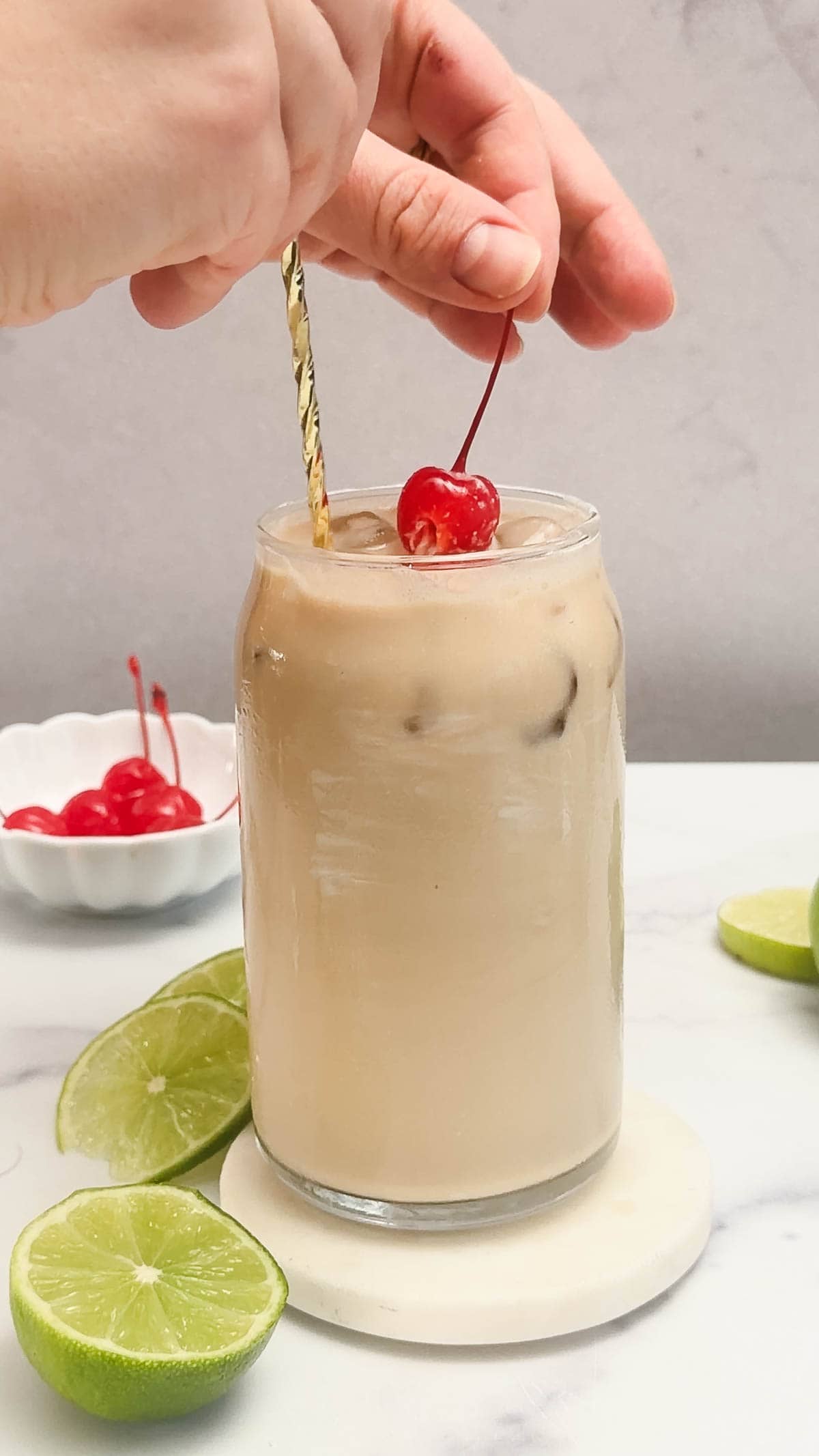 A hand garnishes an iced creamy coconut drink with a cherry; lime slices and cherries are nearby, perfect for a refreshing mocktail recipe.