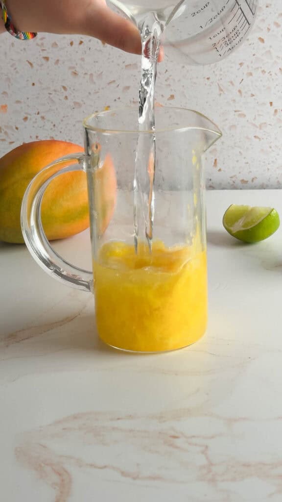Water is being poured into a glass pitcher containing mango pulp, creating a refreshing mango mocktail, with a mango and lime nearby.