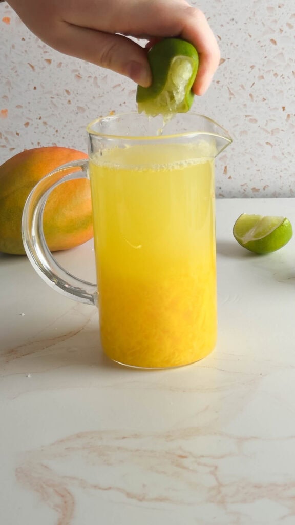 A hand squeezes lime into a glass pitcher of mango juice on a white counter, creating a refreshing Mango Mocktail.