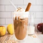 Iced coffee with whipped cream, a glass straw, and cinnamon, inspired by the Apple Crisp Oat Milk Latte&mdash;surrounded by apples, oats, and milk for the perfect Starbucks copycat treat.