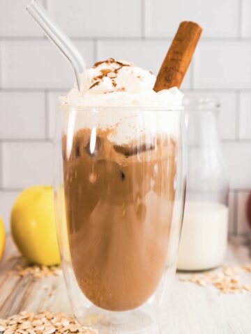 Iced coffee with whipped cream, a glass straw, and cinnamon, inspired by the Apple Crisp Oat Milk Latte&mdash;surrounded by apples, oats, and milk for the perfect Starbucks copycat treat.