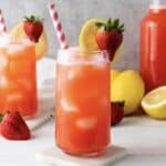 A glass of homemade strawberry lemonade with ice, a straw, lemon slice, and strawberry garnish sits surrounded by fresh fruit.
