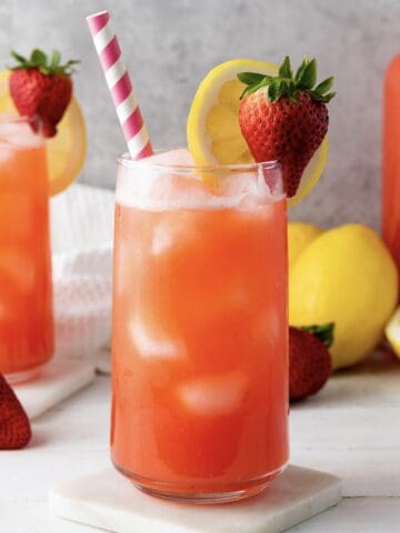 A glass of homemade strawberry lemonade with ice, a straw, lemon slice, and strawberry garnish sits surrounded by fresh fruit.