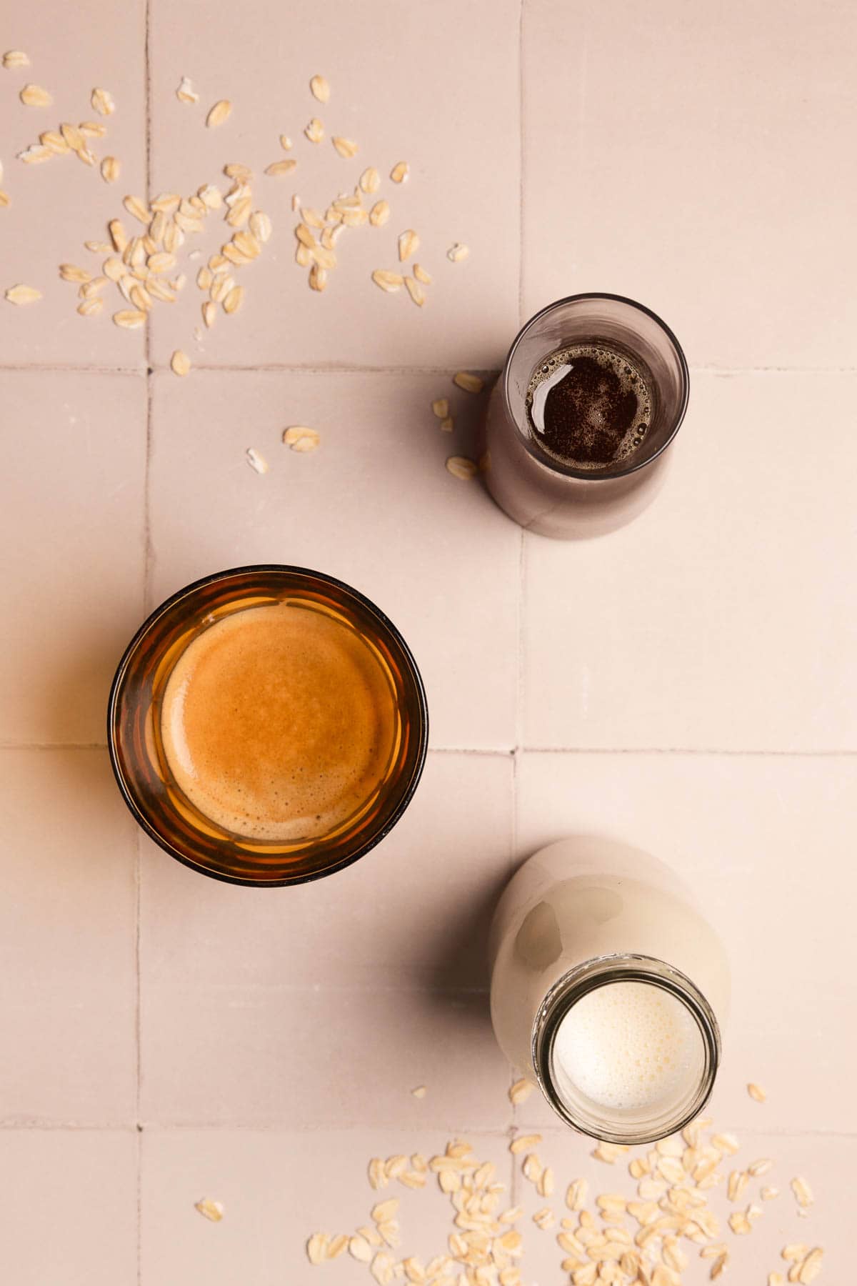Three glass jars filled with coffee, chocolate milk, and oat milk&mdash;perfect for making your own Apple Crisp Oat Milk Latte or Starbucks Copycat drinks&mdash;sit on a tiled surface with scattered oats.