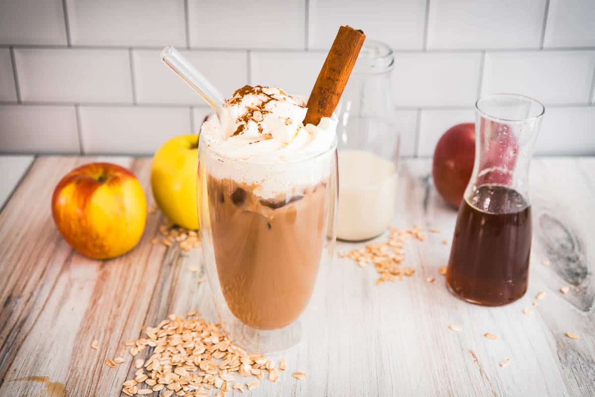 Iced coffee with whipped cream and a cinnamon stick, surrounded by apples, oats, milk, and syrup&mdash;just like a Starbucks Copycat Apple Crisp Oat Milk Latte you can enjoy at home.