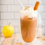 Iced coffee with whipped cream, a cinnamon stick, and a glass straw, next to an apple and scattered oats—just like a homemade Apple Crisp Oat Milk Latte, perfect for a Starbucks Copycat treat.