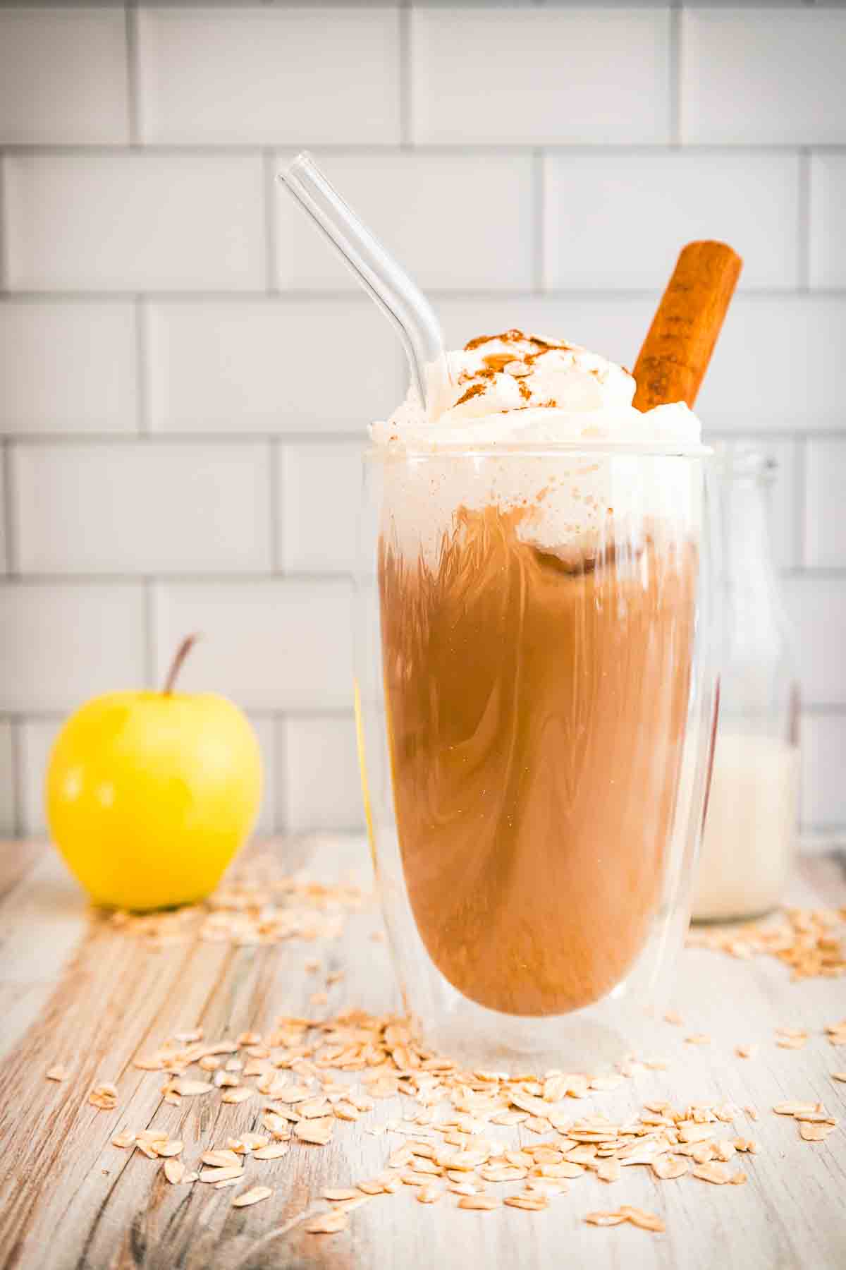 Iced coffee with whipped cream, a cinnamon stick, and a glass straw, next to an apple and scattered oats&mdash;just like a homemade Apple Crisp Oat Milk Latte, perfect for a Starbucks Copycat treat.