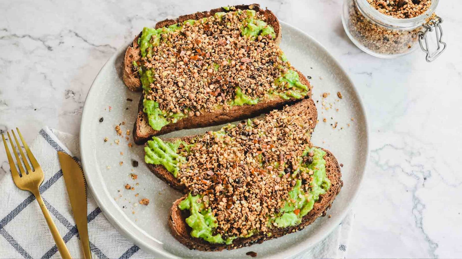Two slices of toast with mashed avocado and a generous sprinkle of seeds on a plate, next to gold utensils.