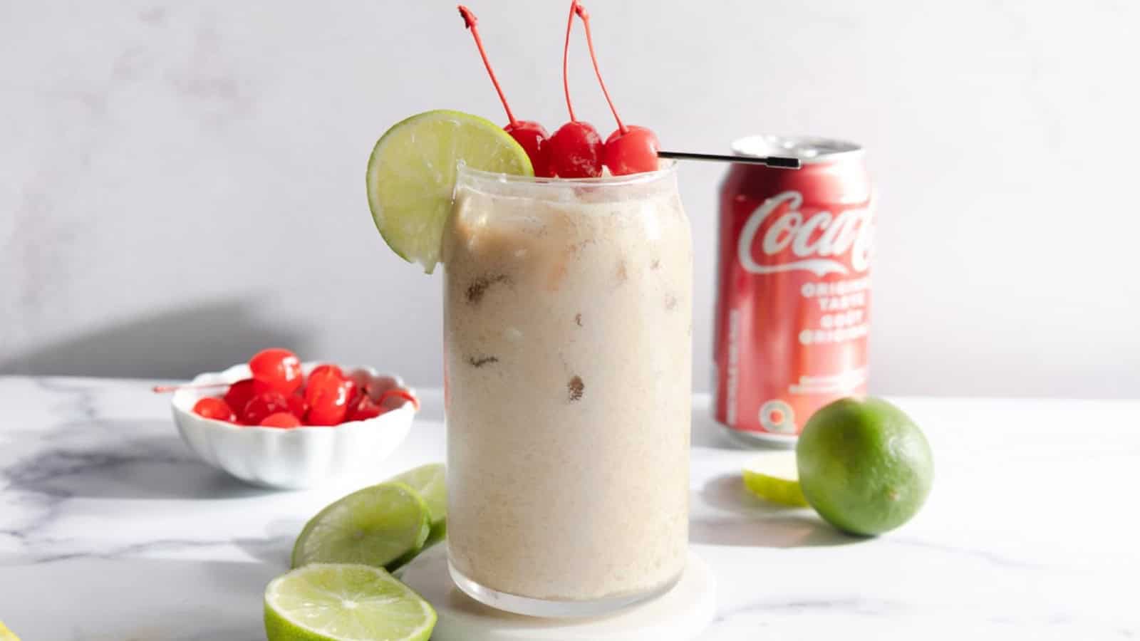 A creamy soda float with cherries and a lime slice in a glass, with a Coke can and limes in the background.