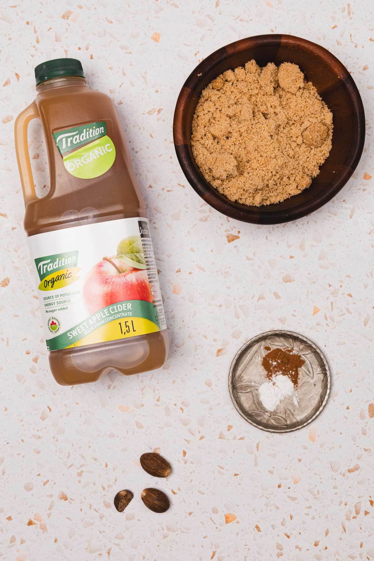 A jug of apple cider, a bowl of brown sugar, spices, and two whole nutmegs on a white speckled surface&mdash;perfect for making a Brown Sugar Apple Crisp Syrup Recipe inspired by your favorite Starbucks copycat drinks.