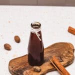A glass bottle of Brown Sugar Apple Crisp Syrup sits on a wooden board, surrounded by whole nutmegs and cinnamon sticks—a perfect Starbucks Copycat syrup recipe for your homemade drinks.