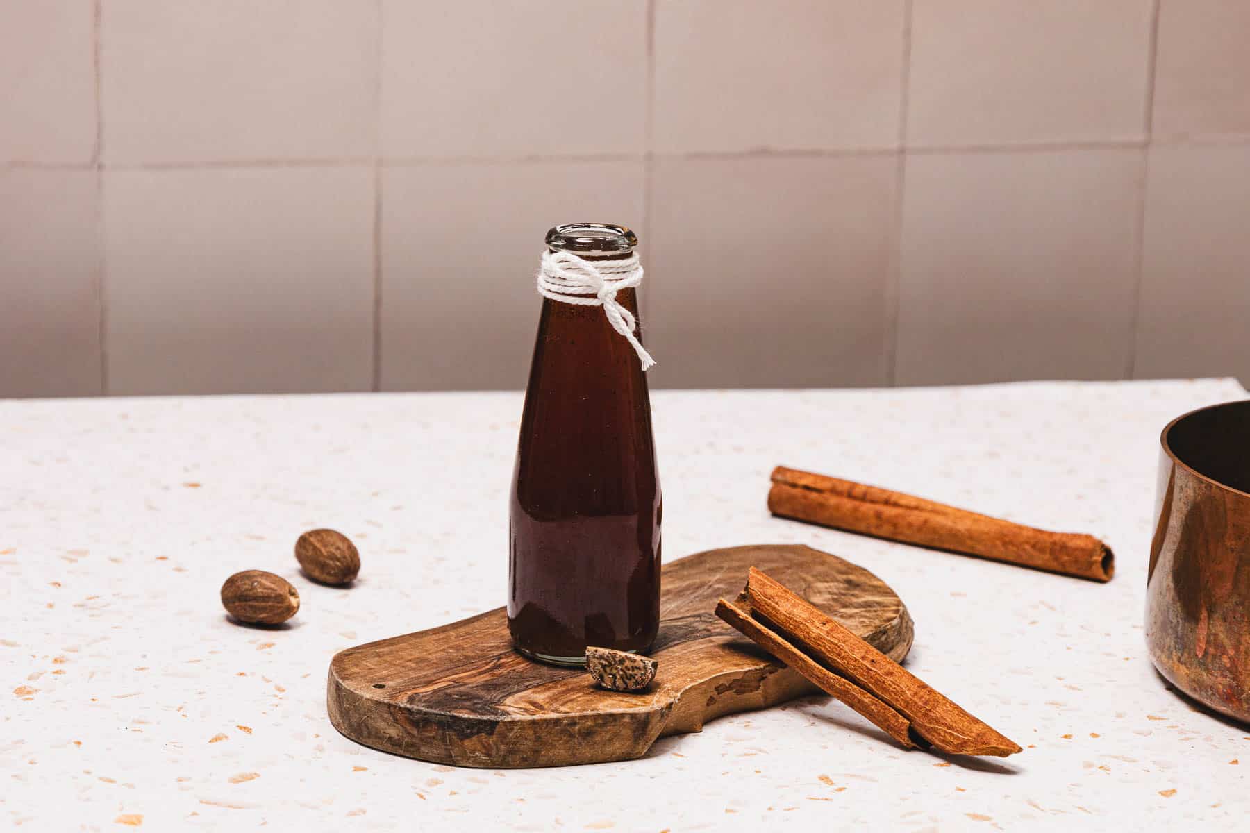 A glass bottle of Brown Sugar Apple Crisp Syrup with twine, spices, and a saucepan on a wooden board atop a white surface&mdash;perfect for your next Starbucks copycat creation.