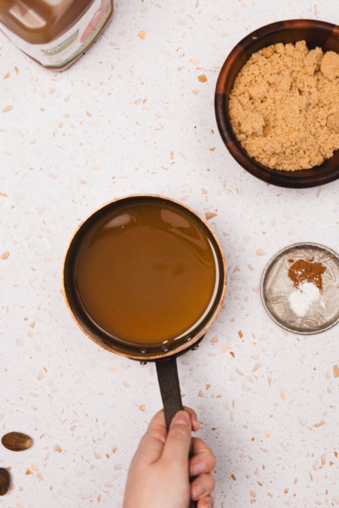 A hand holds a saucepan of homemade Apple Crisp Syrup next to bowls of brown sugar and spices on a white surface, perfect for your next Starbucks copycat creation.