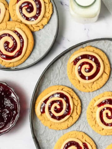 Plates of crumbl copycat peanut butter and jelly cookies with red jam and icing swirls, a bowl of jam, and a bottle of milk on a marble surface.