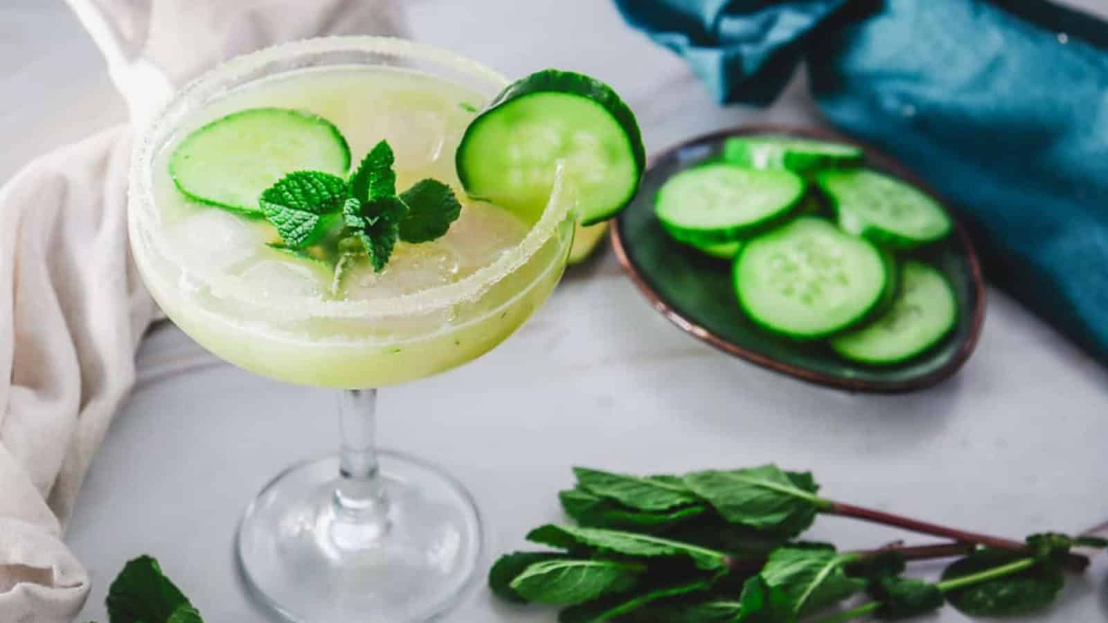 A cocktail garnished with cucumber slices and mint, with more cucumber and mint nearby on a table.