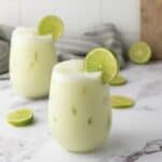 A creamy Brazilian Limeade iced drink in a glass, garnished with a lime slice, sits on a marble surface with additional lime slices nearby.