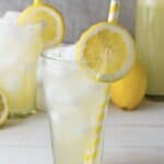 A glass of iced lemonade with a lemon slice and yellow striped straw, surrounded by lemons and more lemonade&mdash;perfect for trying a refreshing Homemade Lemonade Recipe.