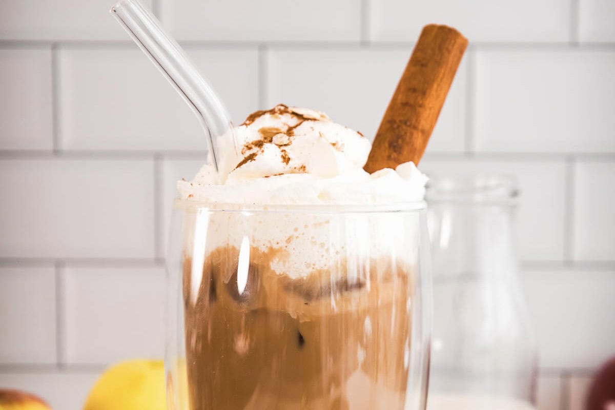 Iced coffee with whipped cream, a cinnamon stick, and a glass straw in front of a white tiled background&mdash;perfect for those craving an Apple Crisp Oat Milk Latte or seeking a Starbucks Copycat experience at home.