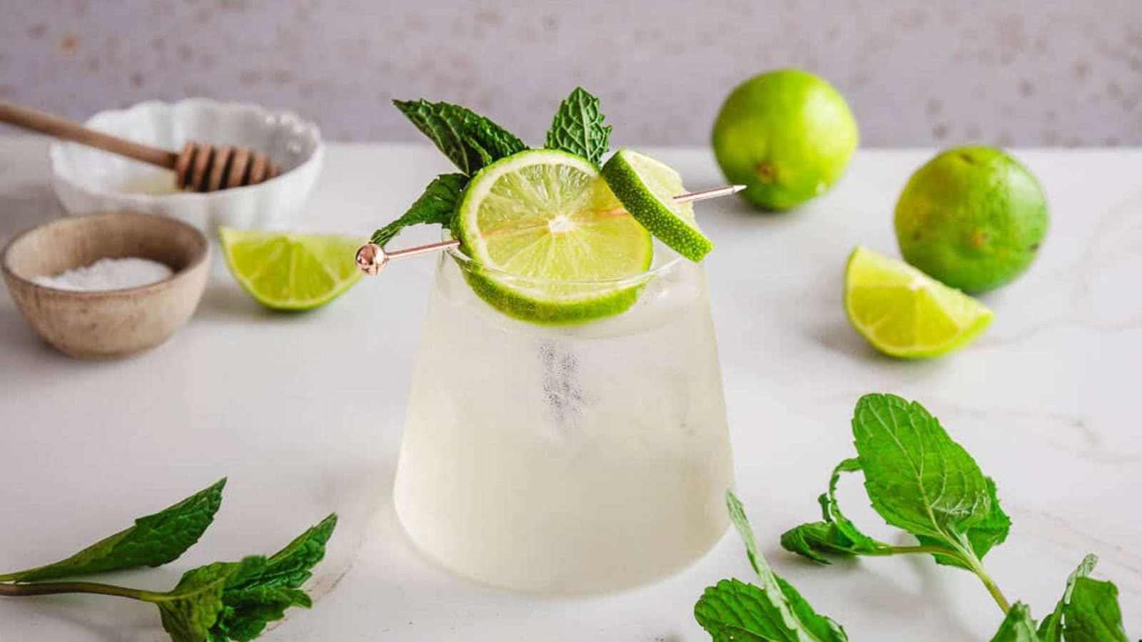 A cocktail garnished with lime and mint on a white surface with fresh limes and mint around it.