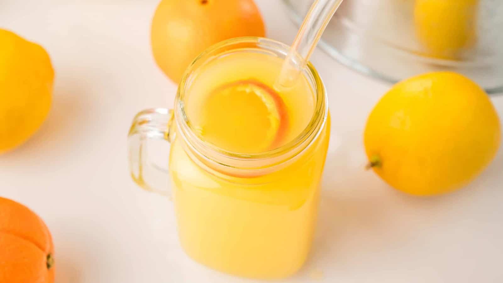 A glass jar of orange juice with a straw and slice of orange, surrounded by fresh citrus fruits.