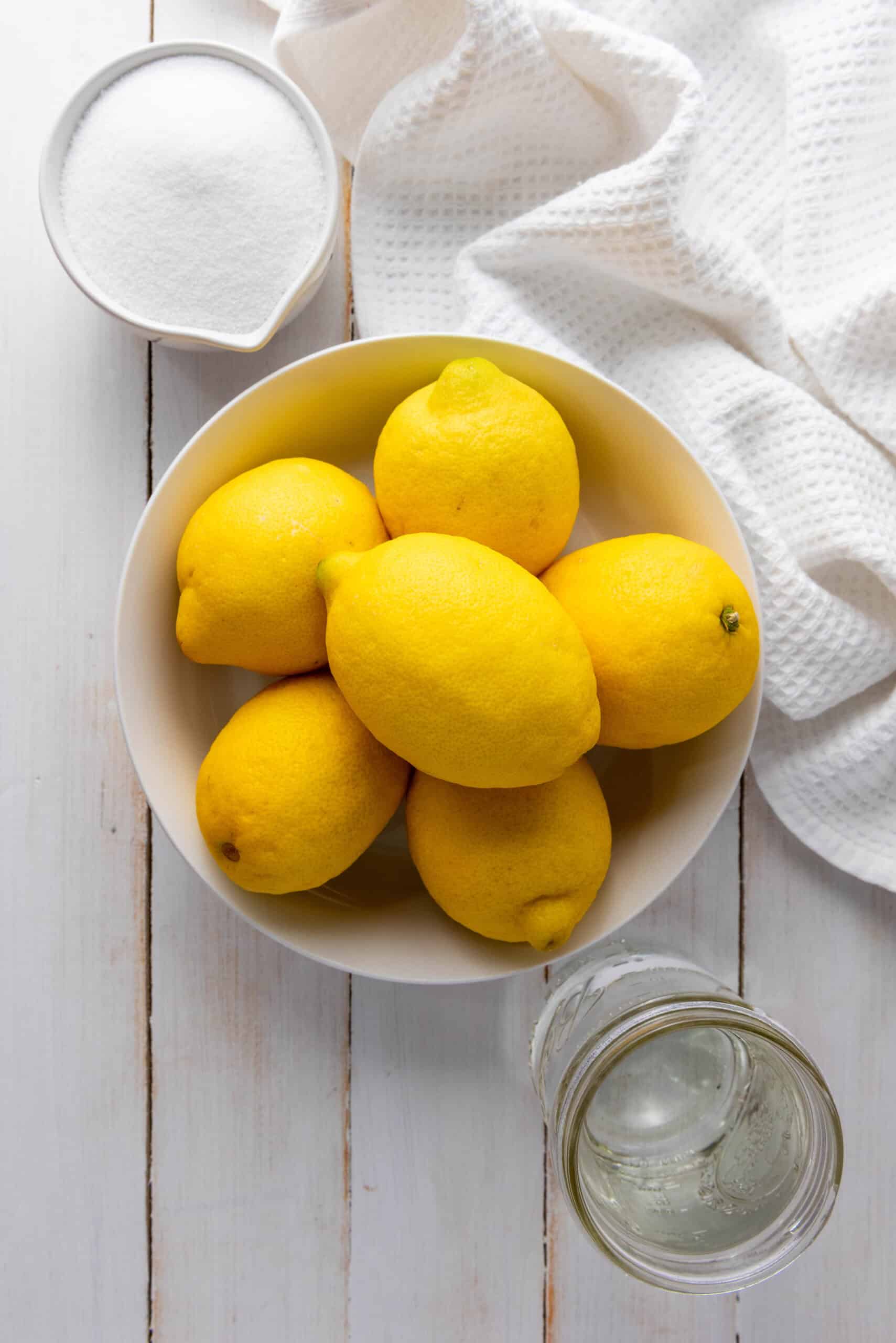 A bowl of lemons, a jar of water, a bowl of sugar, and a white towel on a white wooden surface&mdash;perfect ingredients for trying your favorite Homemade Lemonade Recipe.