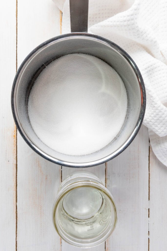 A pot of sugar beside a glass jar and a white towel on a white wooden surface, shot from above&mdash;perfect ingredients for your favorite Homemade Lemonade Recipe.