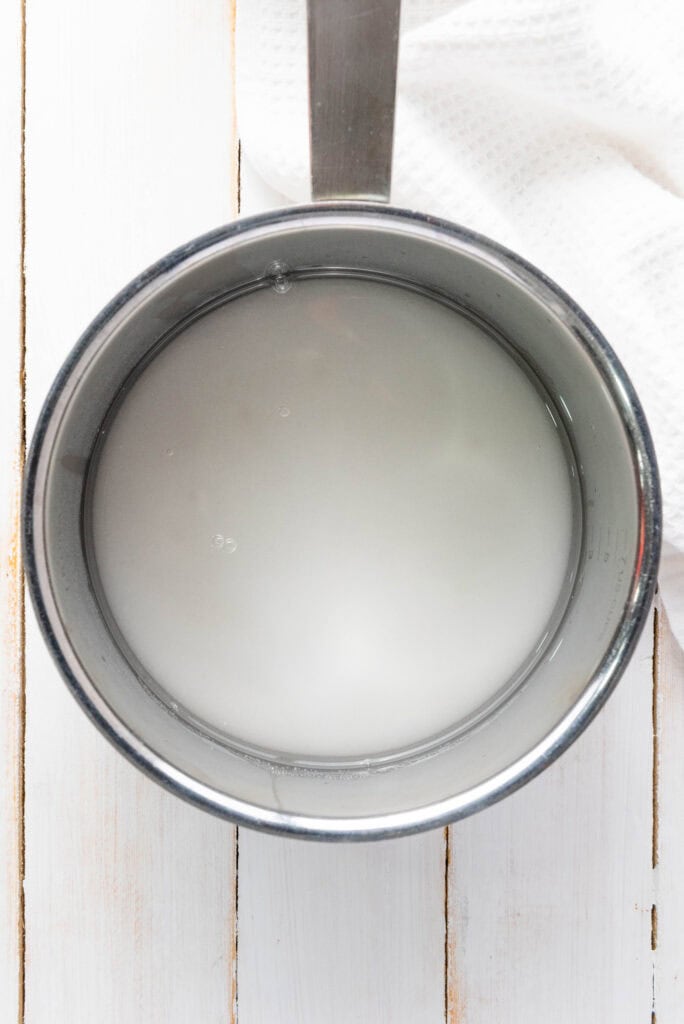 A stainless steel pot with cloudy water, perfect for starting a Homemade Lemonade Recipe, sits on a white wooden surface next to a textured white cloth.