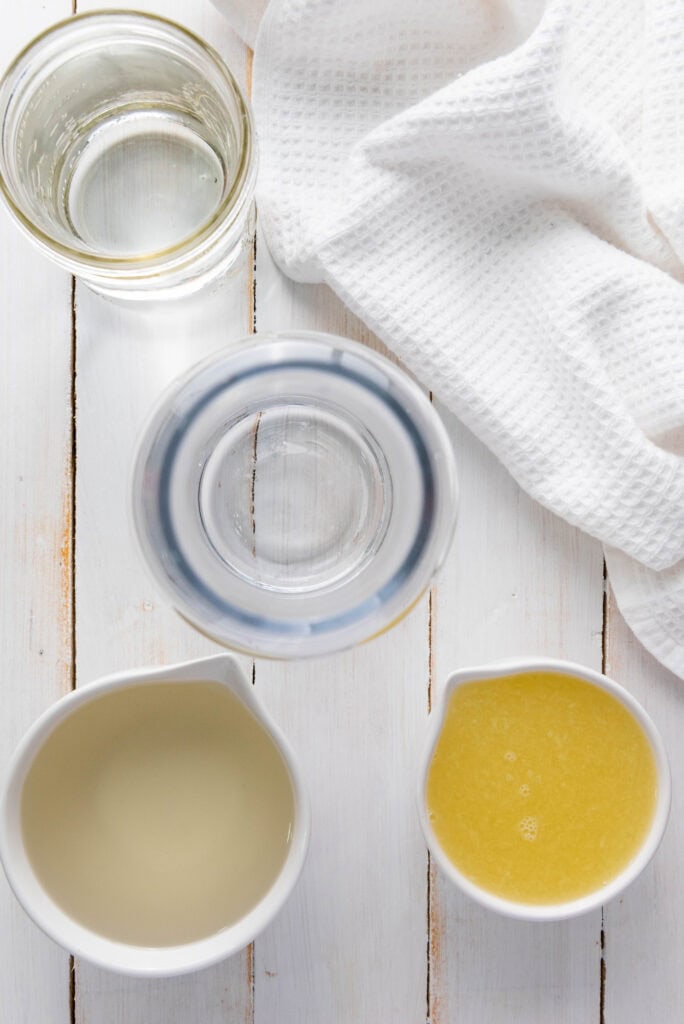 Two small white pitchers with yellow and pale liquid&mdash;perfect for your Homemade Lemonade Recipe&mdash;stand beside a glass of water and a white cloth on a white wooden table.