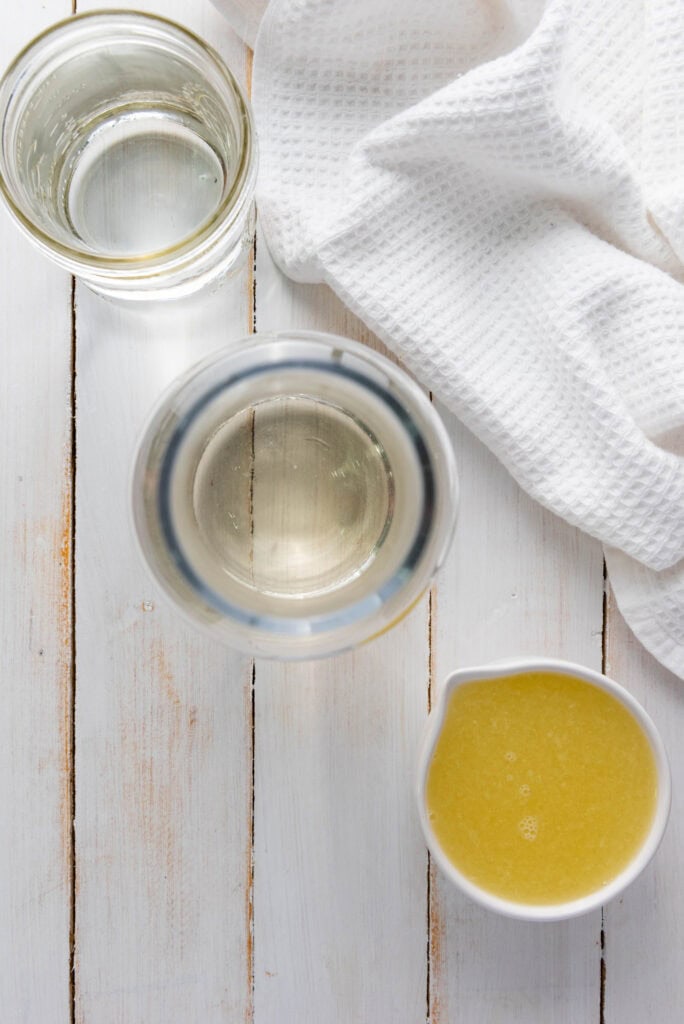 A glass of water, a jug, a cup of yellow liquid reminiscent of homemade lemonade, and a white towel on a white wooden surface.